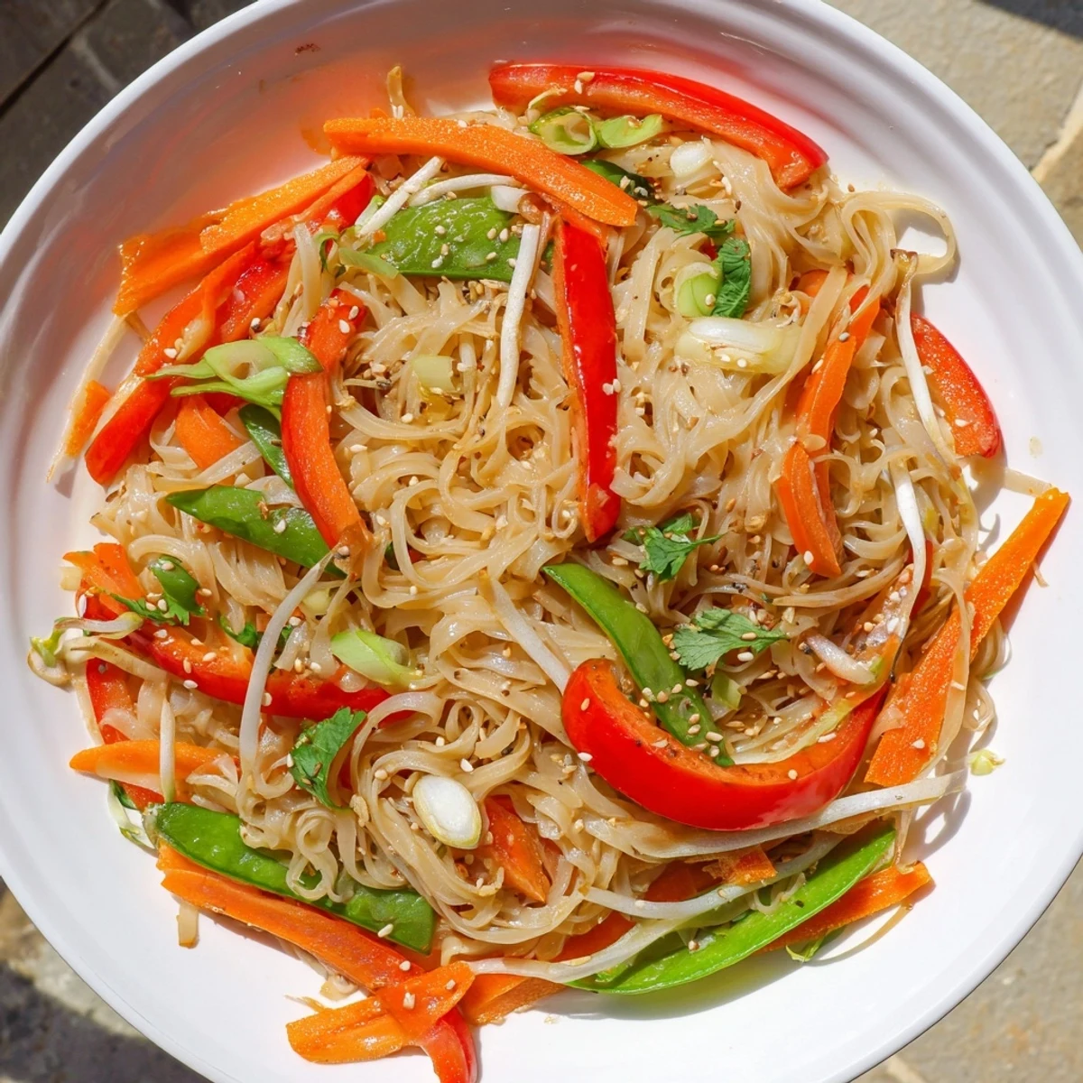 A steaming bowl of Rice Noodle Stir Fry features vibrant carrot ribbons, snap peas, and fresh cilantro garnish.  