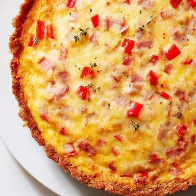 On a white plate, Red Capsicum and Ham Quiche with Sweet Potato Crust is served beside fresh greens for a savory gluten-free dinner.